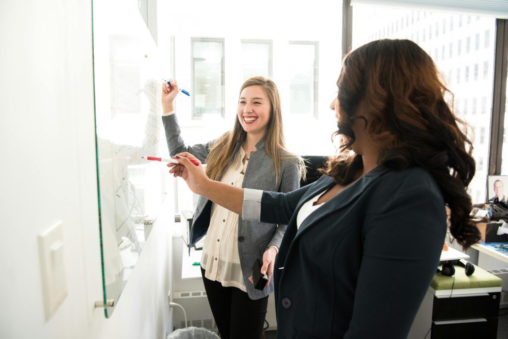 two smiling woman writing on a whiteboard in an office