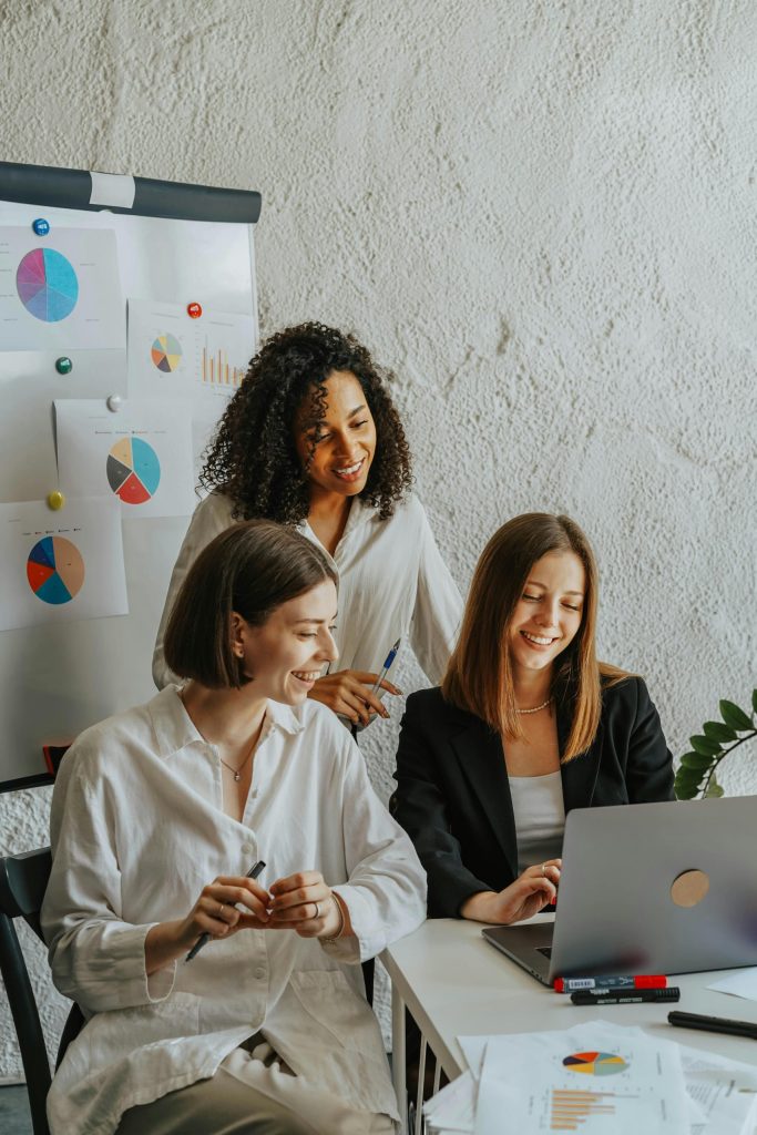 three women smiling looking at a laptop with charts behind them